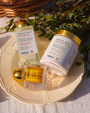 The Vanilla Baby Luxury Trio by Truly Beauty—a body oil, body butter, and shimmer polish—rests on a cream plate with greenery and sunlight, capturing a warm vanilla scent and the promise of glowing skin.