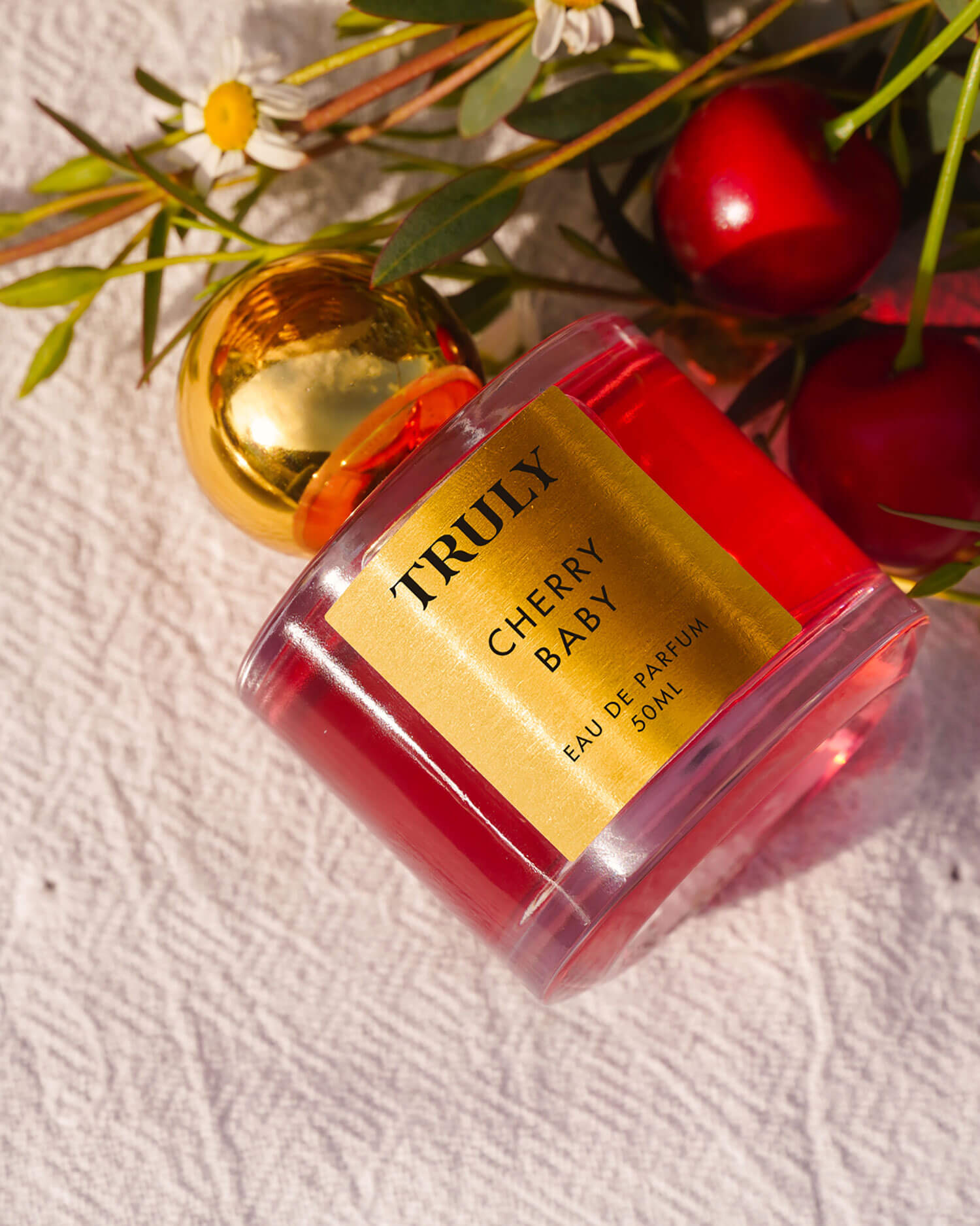 A bottle of Truly Beautys Cherry Baby Eau De Parfum with a gold cap rests on a white textured surface, surrounded by black cherries and green foliage, embodying the lush cherry and delicate jasmine notes.