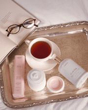 A silver tray holds a cup of tea, a bottle of Truly Beauty Cloud Cream Pro-Peptide Facial Moisturizer, a pink Truly product box, a collapsible silicone cup, an open book, and a pair of eyeglasses.