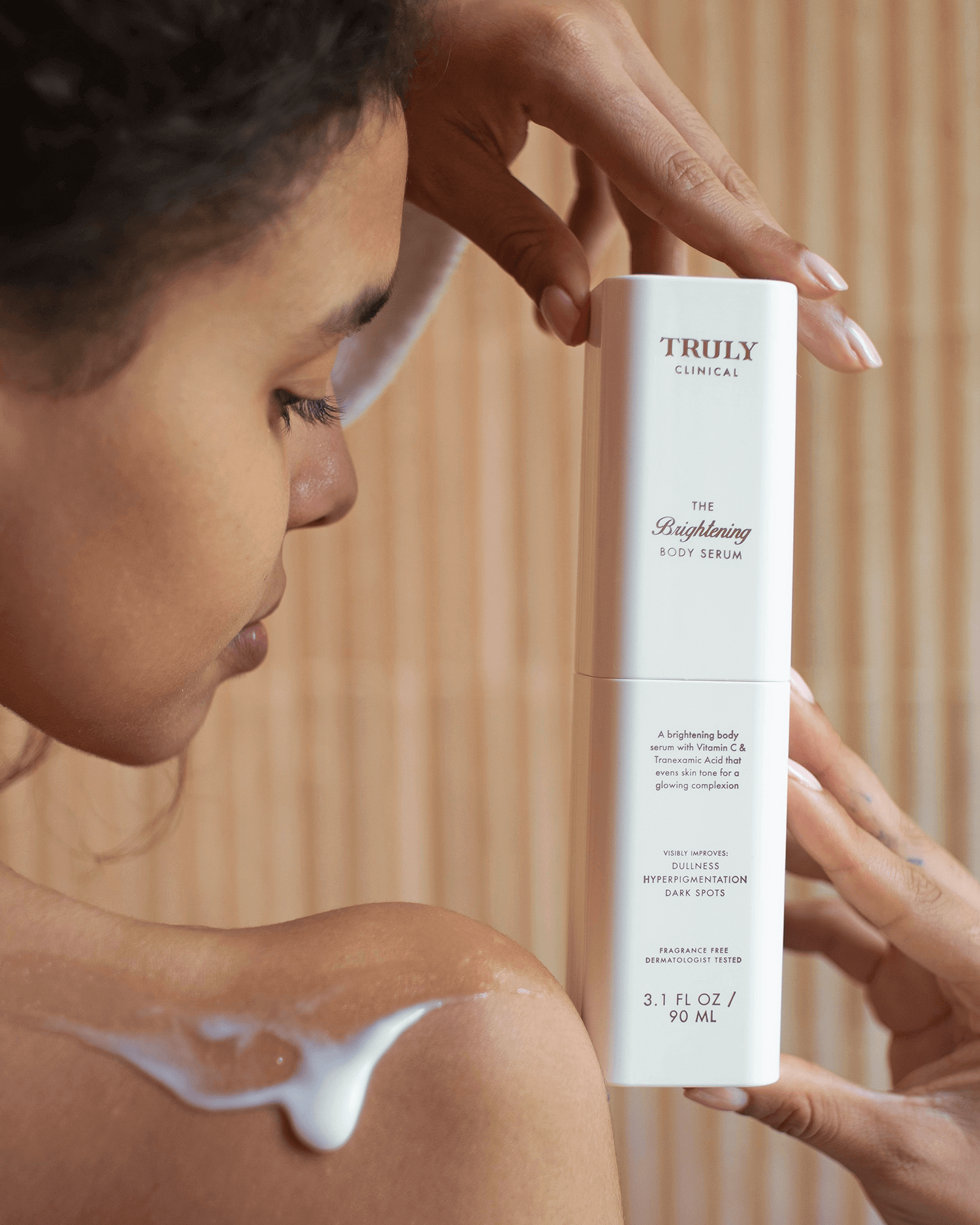 A woman applies a white lotion to her shoulder while holding a bottle of The Body Treatments Complete Collection by Truly Beauty, showcasing effective hydrating body care against a neutral background.