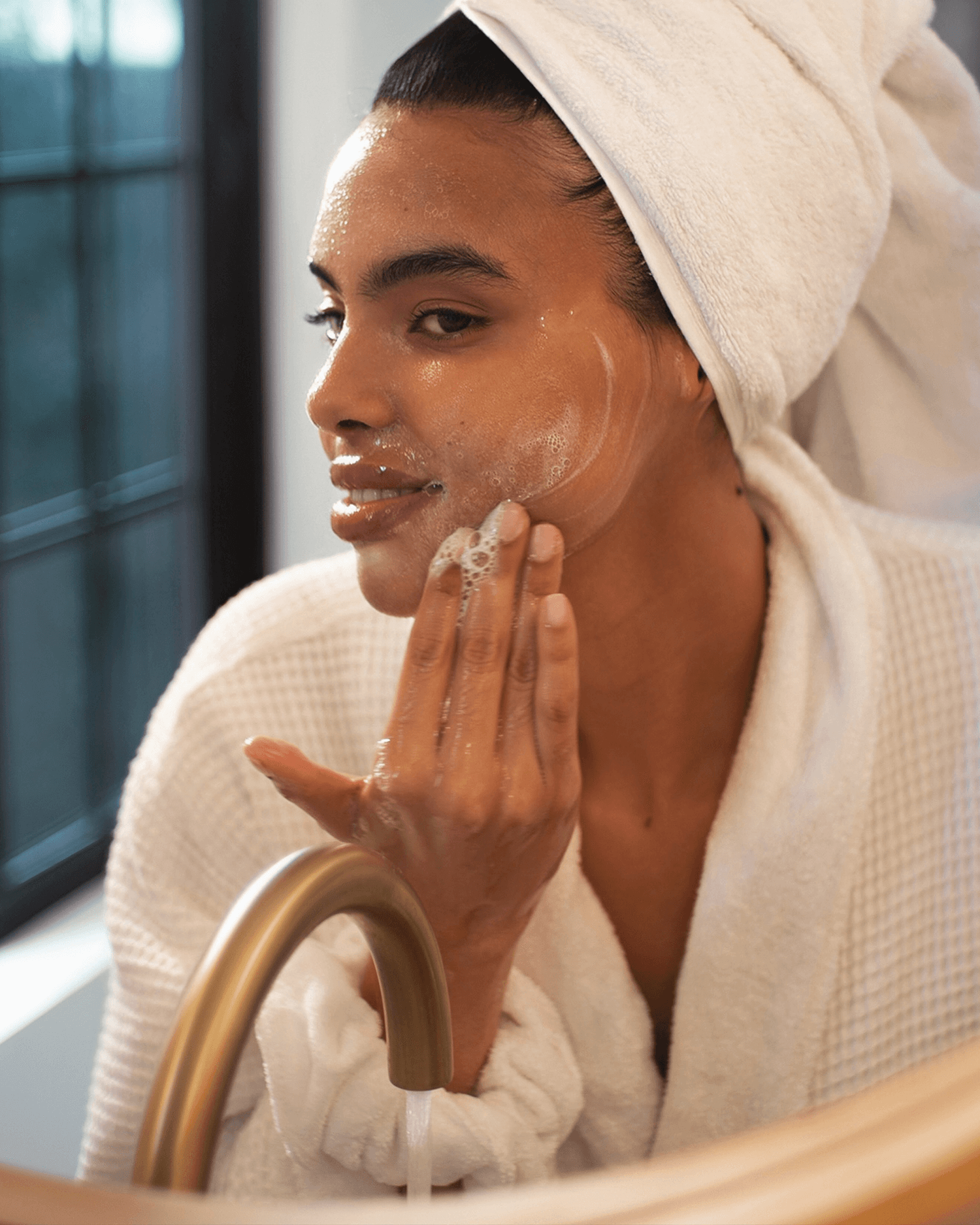 A person in a white bathrobe and towel turban gently cleanses their face at the sink using Truly Beauty's Skin Smoothie Gentle Facial Cleanser.