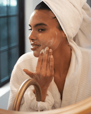 A person in a white bathrobe and towel turban gently cleanses their face at the sink using Truly Beauty's Skin Smoothie Gentle Facial Cleanser.