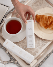 A hand holds a box of The Face Serum Duo by Truly Beauty on a silver tray with tea, a croissant, and another box and bottle of the serum.