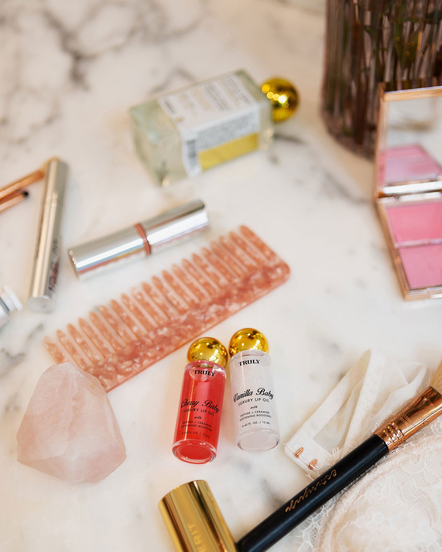 On a marble surface: Truly Beauty’s The Shimmer Duo, plumping lip oil, “Vanilla” and “Cherry” bottles, a pink comb, rose quartz crystal, makeup brush, lipstick, perfume bottle, and blush palette.