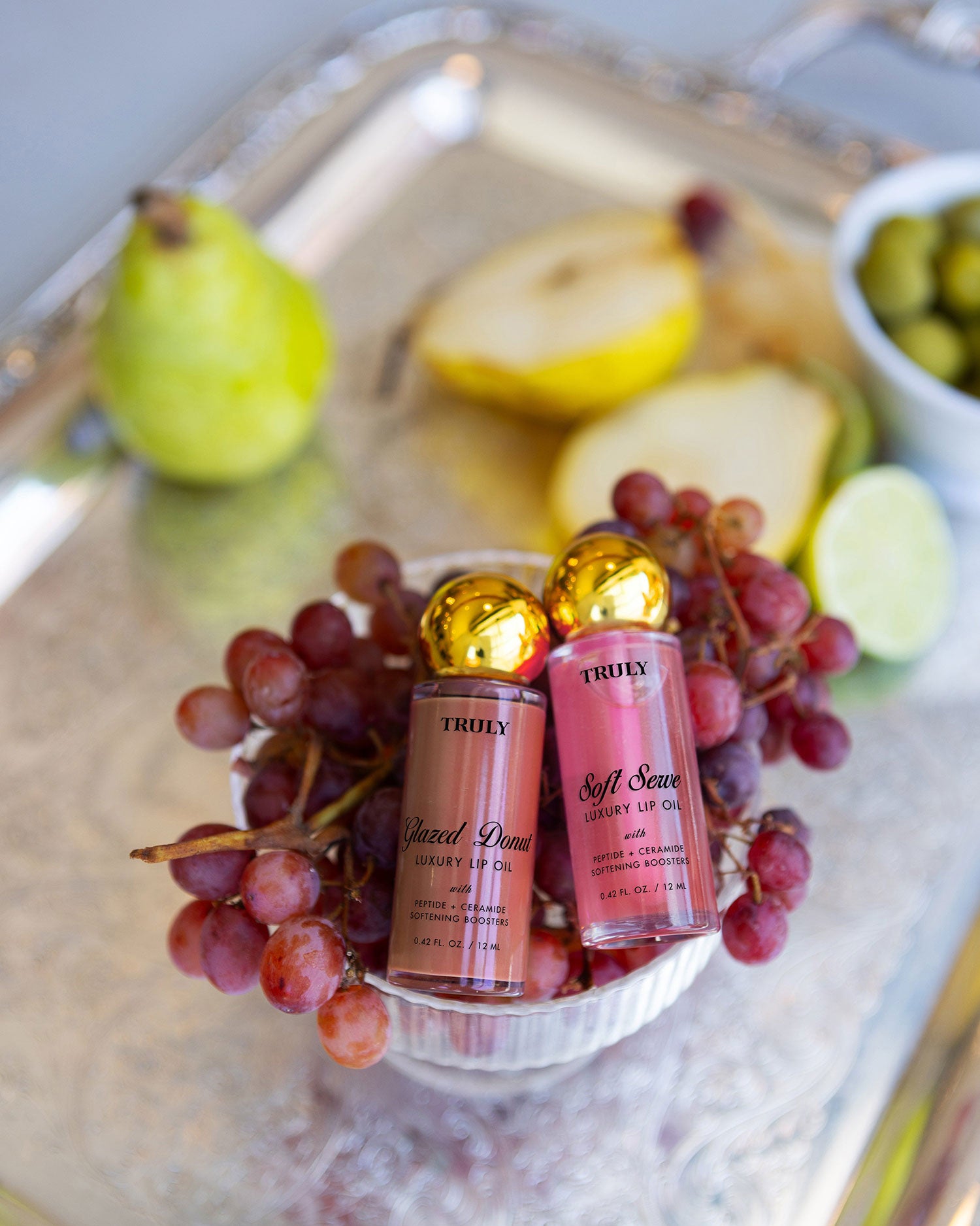 Two tubes of The Glossy Duo by Truly Beauty are nestled among red grapes in a small bowl on a silver tray, with a sliced pear, lime, and green olives in the background.
