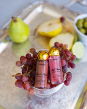 Two tubes of The Glossy Duo by Truly Beauty are nestled among red grapes in a small bowl on a silver tray, with a sliced pear, lime, and green olives in the background.