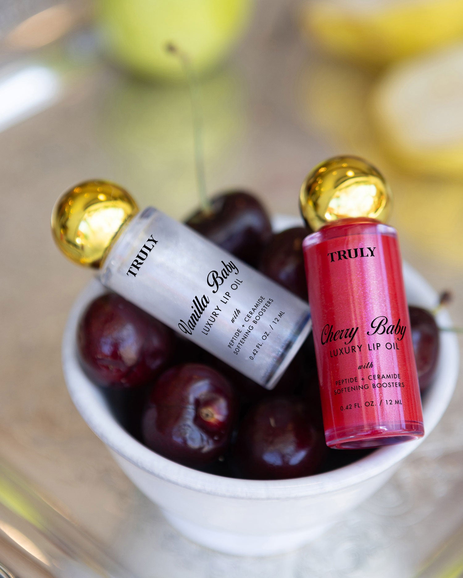 Two bottles of The Shimmer Duo lip oil by Truly Beauty, in Vanilla Baby and Cherry Baby, rest on dark cherries in a white bowl. Their gold caps pop against a blurred backdrop with flashes of yellow.