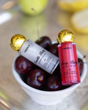 Two bottles of The Shimmer Duo lip oil by Truly Beauty, in Vanilla Baby and Cherry Baby, rest on dark cherries in a white bowl. Their gold caps pop against a blurred backdrop with flashes of yellow.