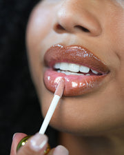 A close-up of a woman applying Truly Beauty’s The Glossy Duo to her lips with an applicator wand, revealing smooth skin, glossy makeup, and shiny nude hydration.