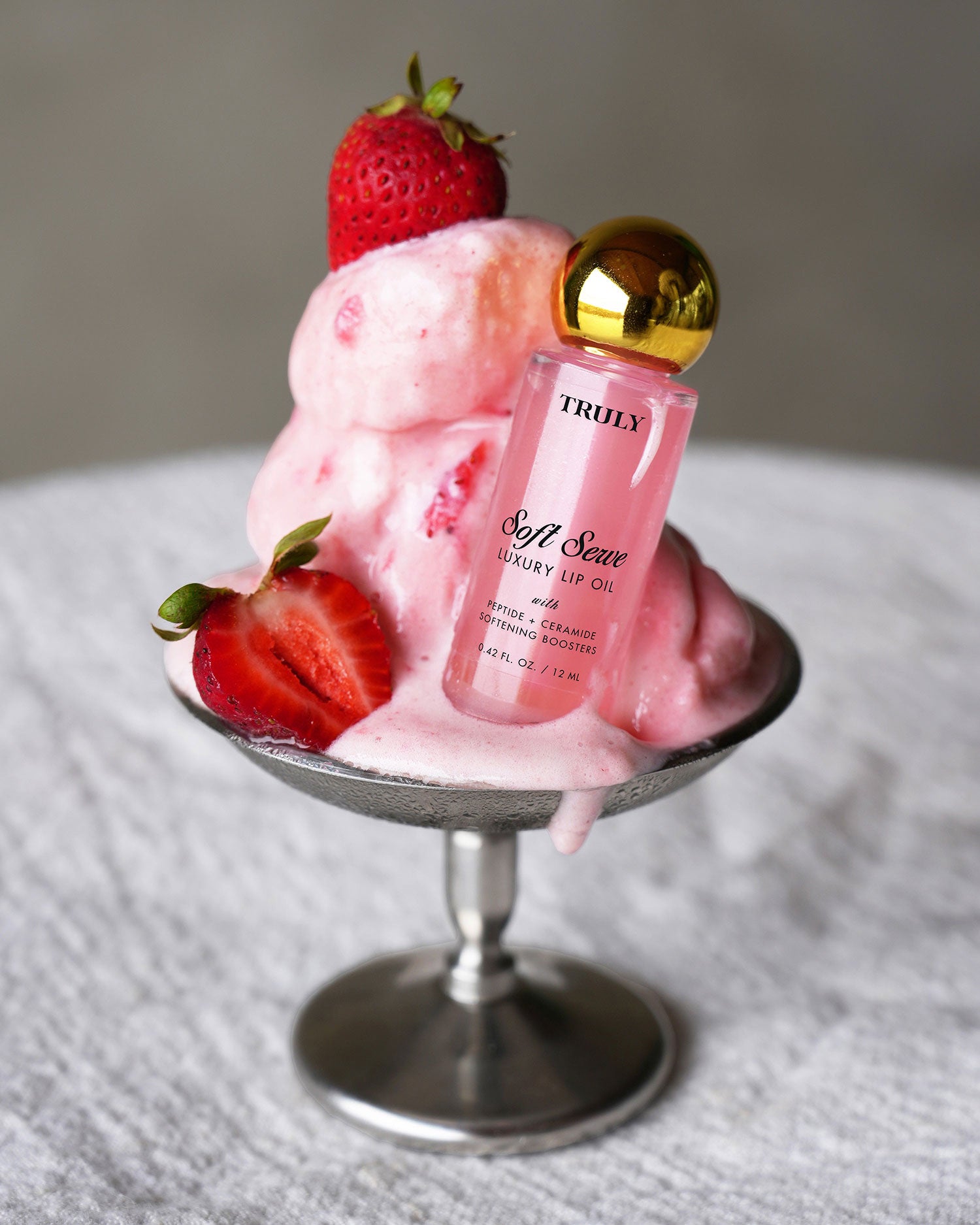 The Glossy Duo by Truly Beauty sits beside melting pink ice cream and fresh strawberries in a glass dish, all arranged on a white textured surface.