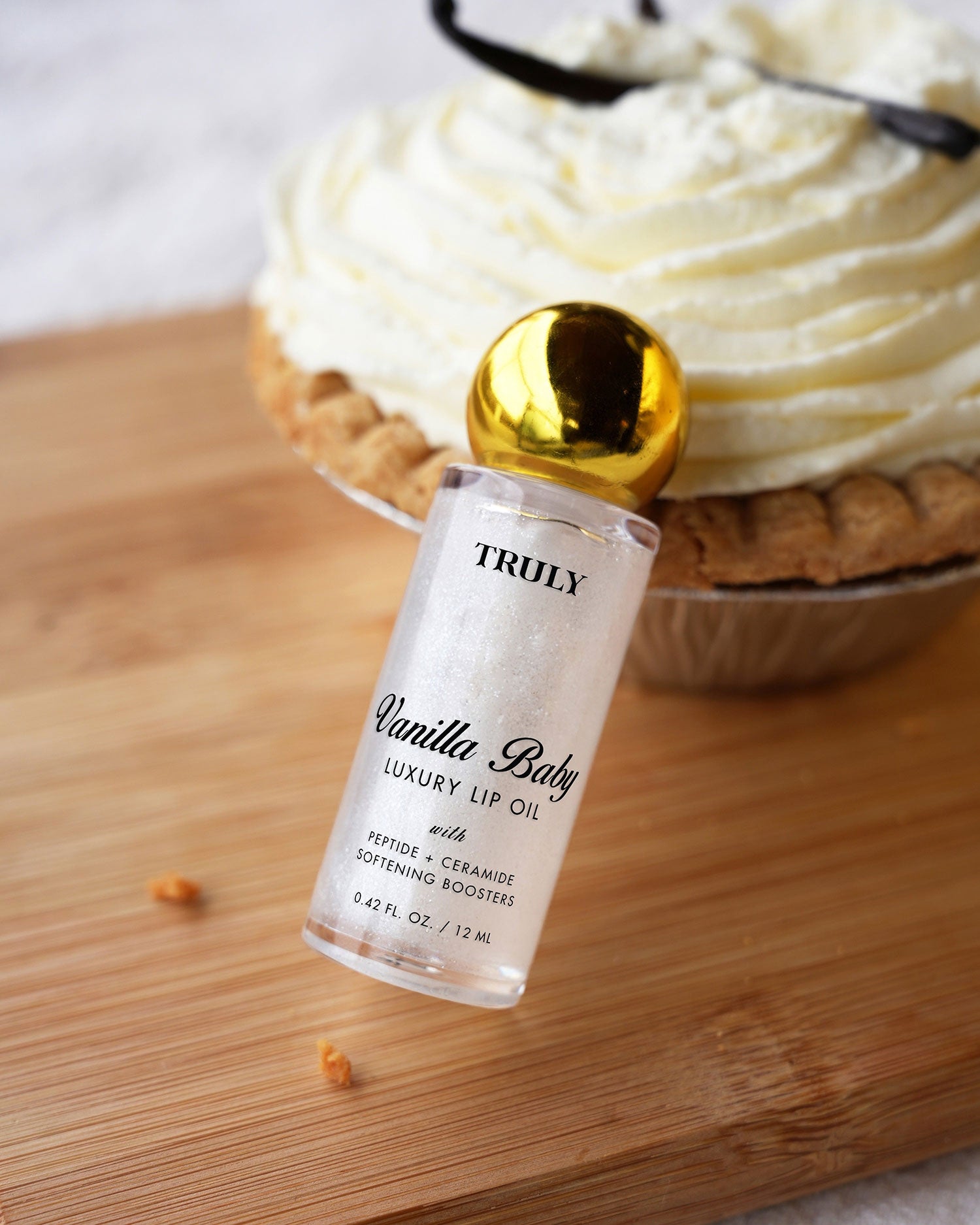 A bottle of Truly Beauty’s The Shimmer Duo with a gold cap stands upright in front of a whipped cream-topped pie on a wooden surface.
