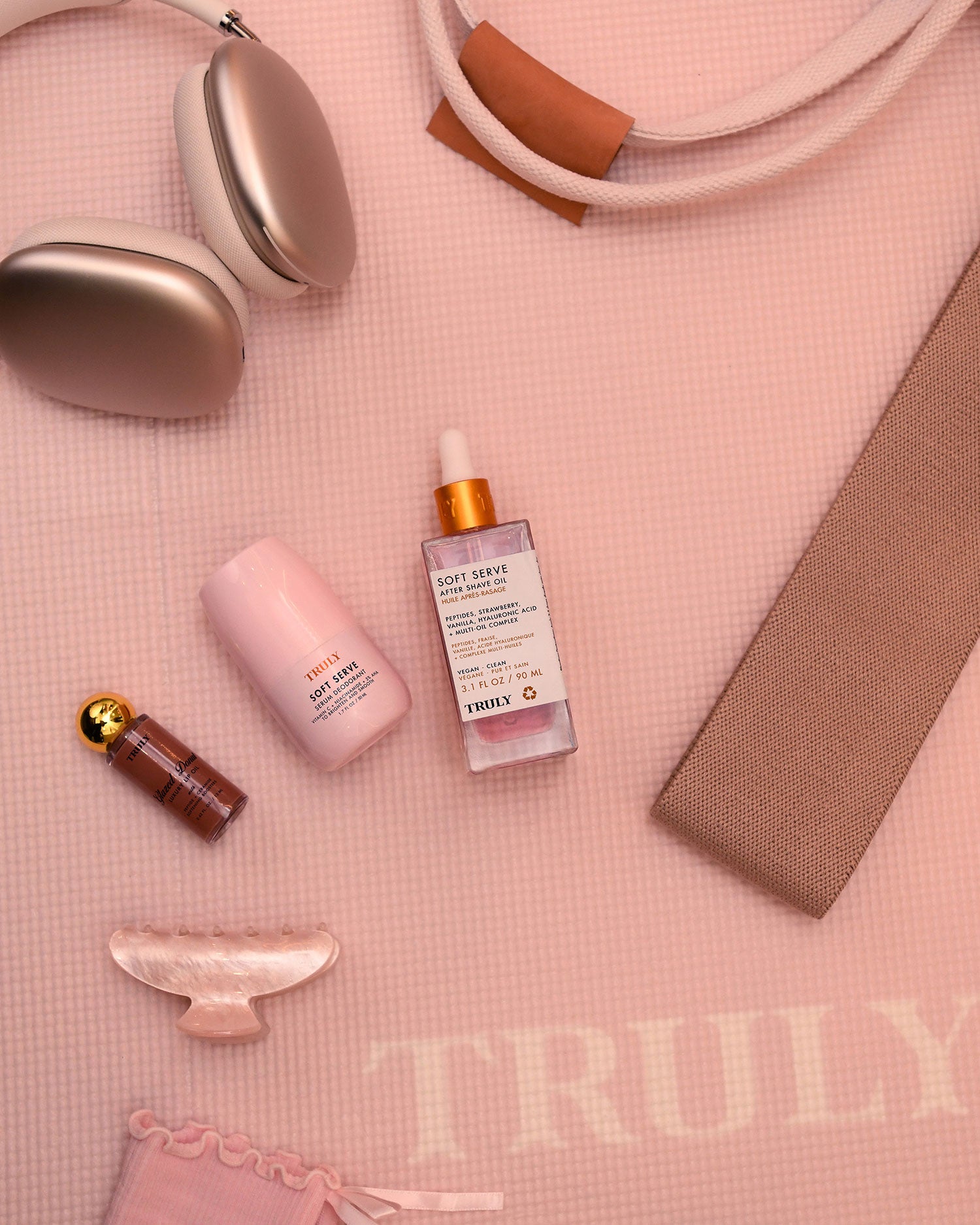 Flat lay of beauty products and accessories on a pale pink mat with TRULY text. Featured items include headphones, Truly Beauty Soft Serve Serum Deodorant, a serum bottle, a pink skincare bottle, lipstick, hair clip, and a beige strap.