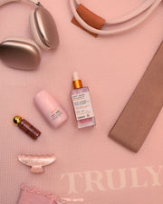 Flat lay of beauty products and accessories on a pale pink mat with TRULY text. Featured items include headphones, Truly Beauty Soft Serve Serum Deodorant, a serum bottle, a pink skincare bottle, lipstick, hair clip, and a beige strap.