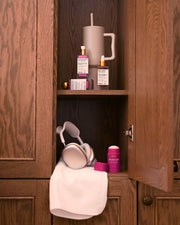 A wooden cabinet with an open door displays Truly Beauty’s The Viral Edit skincare set, a large tumbler with a straw, purple headphones on a white towel, and a pink container of wellness powder.