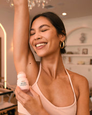 A smiling woman with closed eyes applies Truly Beauty’s Soft Serve Serum Deodorant to her raised armpit, looking happy and relaxed in a bright, stylish room.