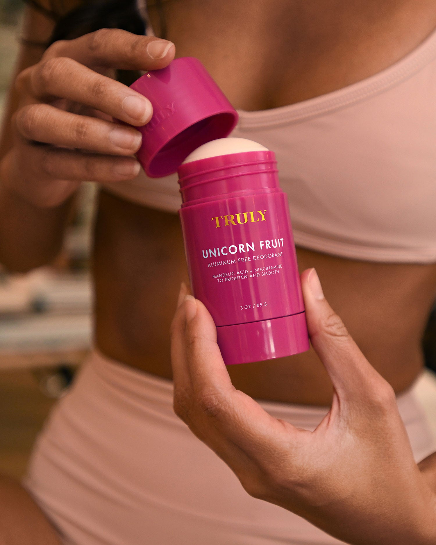 A person in a beige bikini opens a pink stick of Truly Beauty’s The Viral Edit deodorant, holding it close to their body and lifting the lid with the other hand.