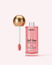 A bottle of Truly Beauty Luxury Peptide Lip Oil with pink liquid and a gold-capped applicator wand dipped in the oil, set against a light pink background.
