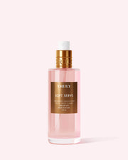 A clear pink bottle of Truly Beauty Soft Serve Perfume Mist with a gold label and cap, boasting a sweet strawberries and cream scent, is photographed against a light pink background.