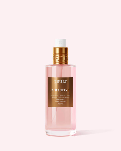 A clear pink bottle of Truly Beauty Soft Serve Perfume Mist with a gold label and cap, boasting a sweet strawberries and cream scent, is photographed against a light pink background.