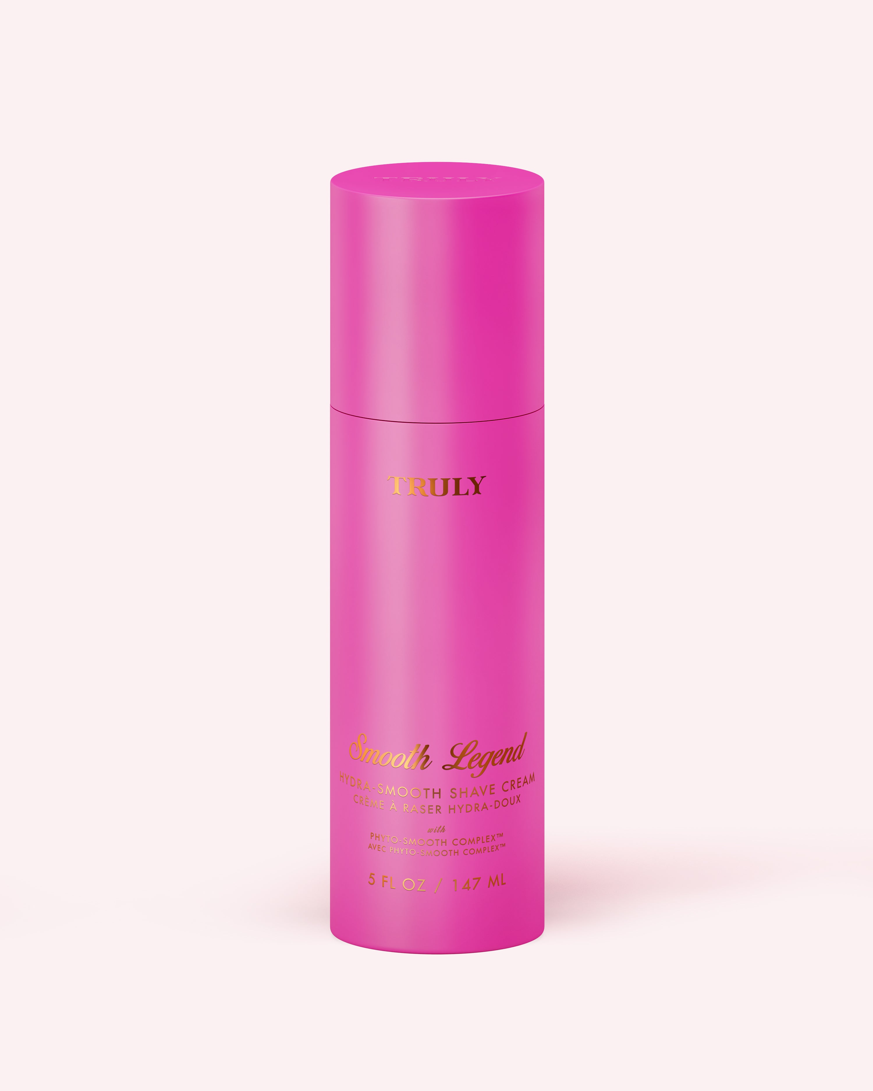 A tall, cylindrical pink bottle labeled Truly Beauty and Hydra-Smooth Shave Cream, with gold text and a matching pink cap, stands upright on a light pink background.