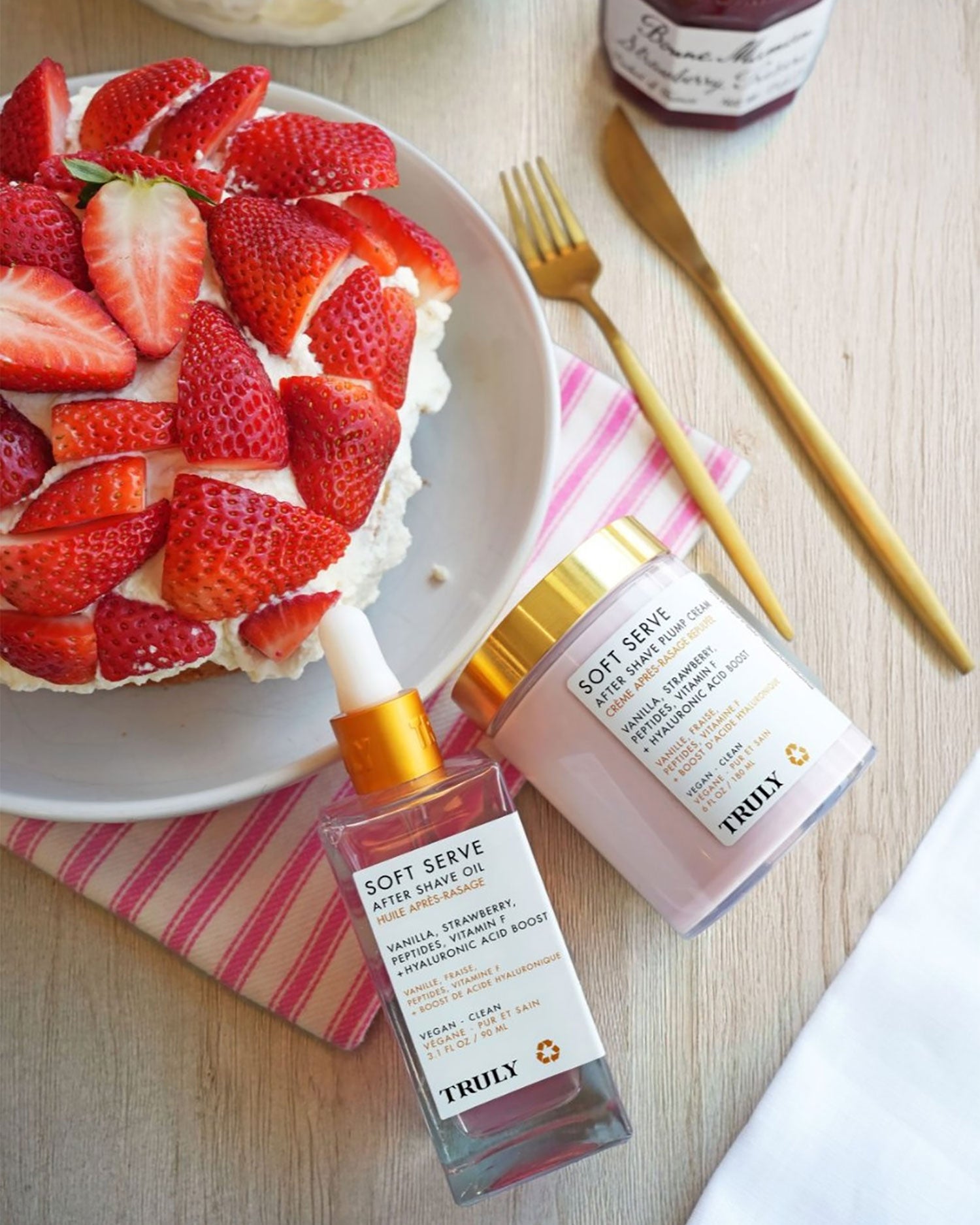 A cake with fresh strawberries sits on a plate beside two Truly Beauty Soft Serve After Shave Duo products—a serum and a cream—on a pink striped cloth with a gold-handled fork nearby.