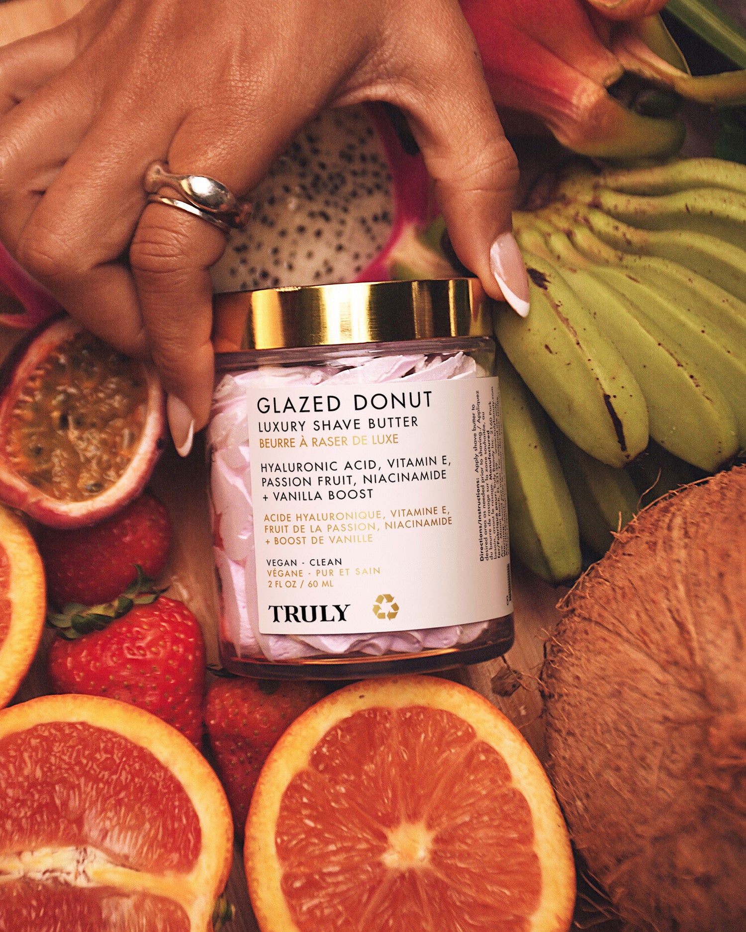 A hand with manicured nails holds a jar of Truly Beauty’s Glazed Donut Luxury Shave Butter, made with hydrating hyaluronic acid, surrounded by sliced citrus fruits, kiwi, and banana on a wooden surface.