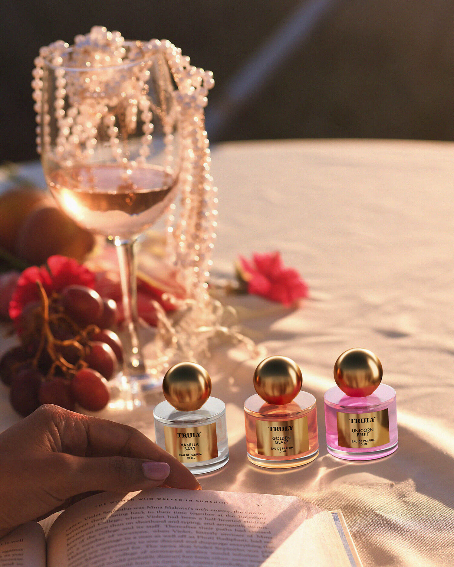 A glass of rosé, a pearl necklace, grapes, and The Perfumer’s Set by Truly Beauty rest on the table. A hand opens a book amid flowers and gentle sunlight, creating a warm, elegant atmosphere.