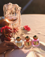 A glass of rosé, a pearl necklace, grapes, and The Perfumer’s Set by Truly Beauty rest on the table. A hand opens a book amid flowers and gentle sunlight, creating a warm, elegant atmosphere.