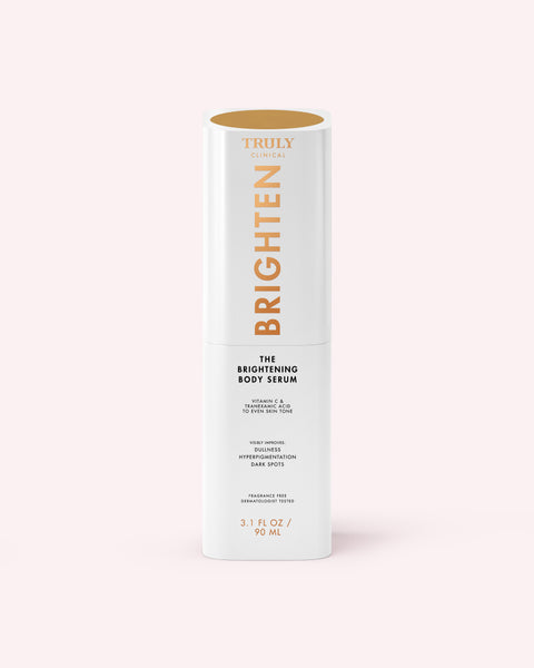 A white cylindrical bottle of The Brightening Body Serum by Truly Beauty, formulated to target dark spots, stands upright with gold and black text on a pale pink background.