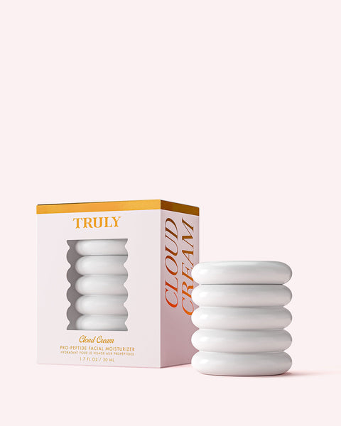 A white, ribbed jar of Truly Beauty’s Cloud Cream Pro-Peptide Facial Moisturizer sits beside its white, gold-accented box with a cutout displaying the jar. Both are set against a pale pink background.