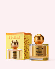A bottle of Truly Beauty Golden Glaze Eau De Parfum with a shiny gold cap sits beside its matching yellow and gold box on a light pink background, evoking the warm essence of guaiacwood oil within.