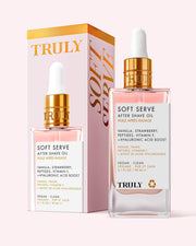 A bottle of Soft Serve After Shave Oil stands before its matching box. The pink, gold, and white packaging showcases details and ingredients like hyaluronic acid and peptides for post-shave skin care.
