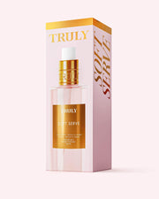 A bottle of Truly Beauty Soft Serve Perfume Mist stands before its matching pink and gold box, both displaying the brand name and product label, evoking a strawberries and cream fragrance with a hint of vanilla.