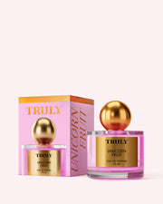 A 50 ml bottle of Truly Beauty Unicorn Fruit Eau De Parfum, featuring a gold label and round gold cap, is displayed beside its pink and orange box with gold lettering. This fragrance has a fruity, feminine scent inspired by candy floss.