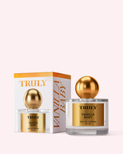 A bottle of Truly Beauty Vanilla Baby Eau De Parfum with a gold cap sits beside its matching white and gold box on a pale pink background, suggesting notes of amber, caramel drizzle, and bold vanilla.
