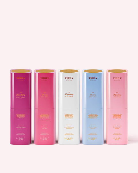 Five colorful cylindrical bottles from Truly Beauty’s The Body Treatments Complete Collection, each with a gold lid, are lined up against a light pink background in shades of pink, magenta, red, white, and light blue.