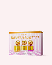 The Perfumers Set by Truly Beauty includes three gold-lidded jars—Golden Glaze, Vanilla Baby, and Unicorn Fruit—each with a unique scent, all displayed on a pink and white striped background.