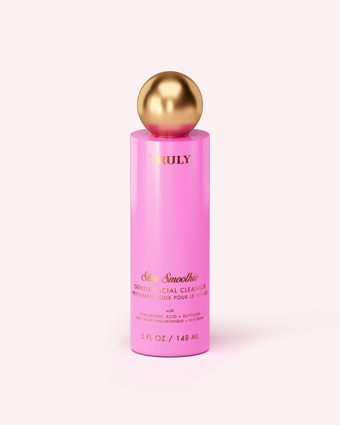 A pink bottle of Truly Beauty’s Skin Smoothie Gentle Facial Cleanser, featuring gold text and a gold spherical cap, sits on a light pink background. The hydrating formula with hyaluronic acid comes in a 5 fl oz (148 mL) size.