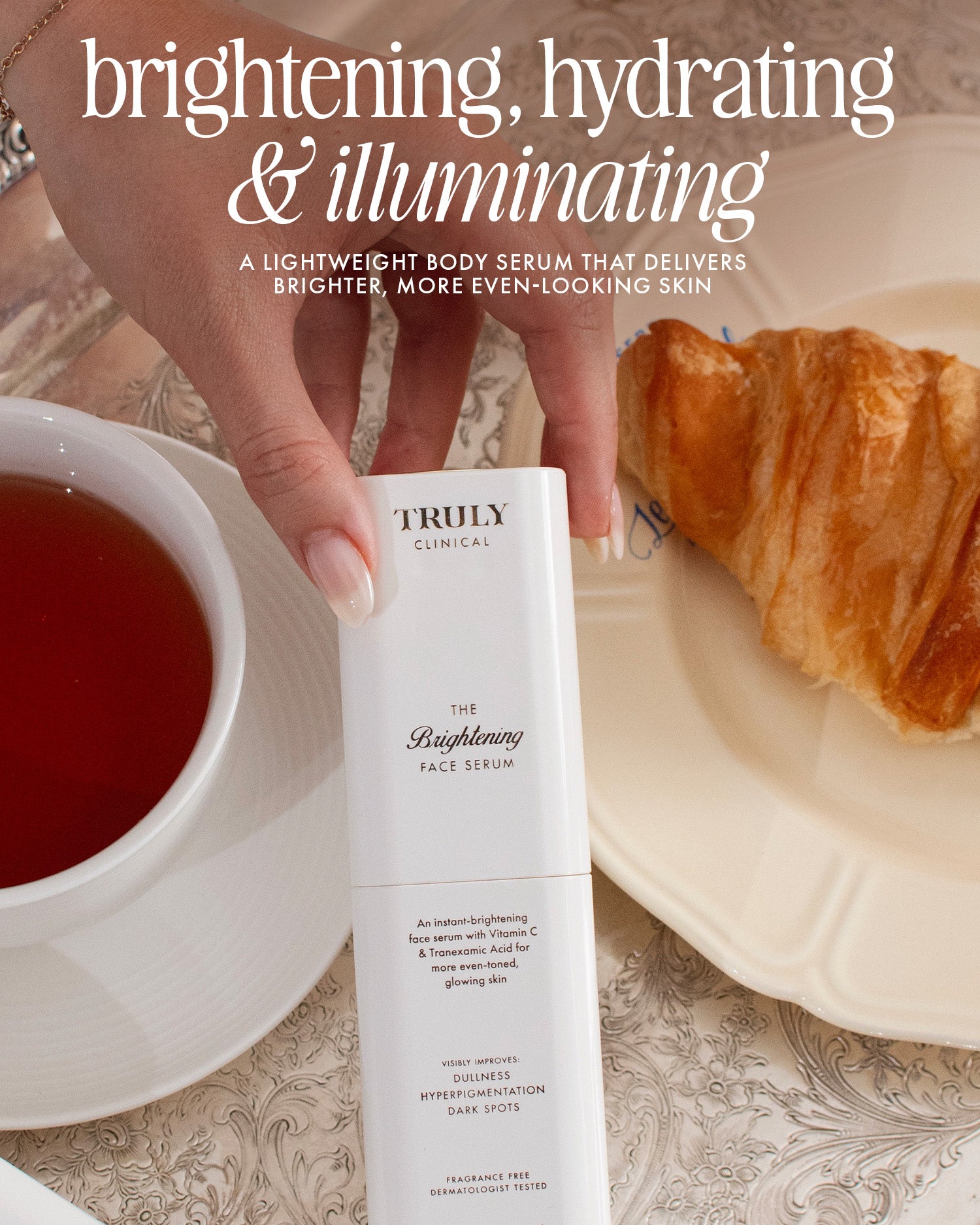 A hand holds a bottle of The Face Serum Duo by Truly Beauty over a table with a red drink, a plate, and a croissant. White text above highlights its brightening, hydrating, and illuminating benefits.