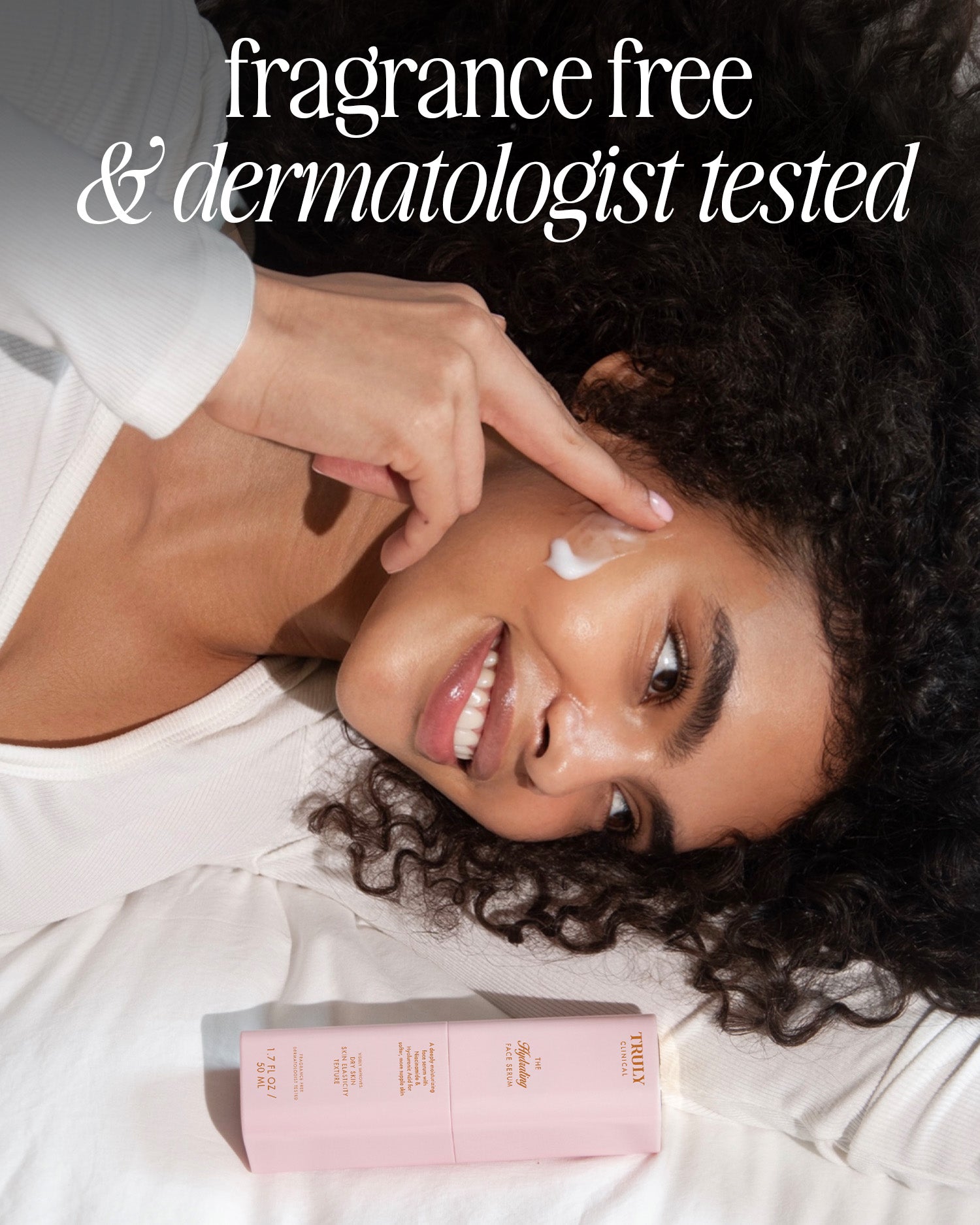 A smiling woman lies on white bedding, applying The Hydrating Face Routine by Truly Beauty to her cheek. Next to her is a pink bottle. The text above reads, “fragrance free & dermatologist tested.”.