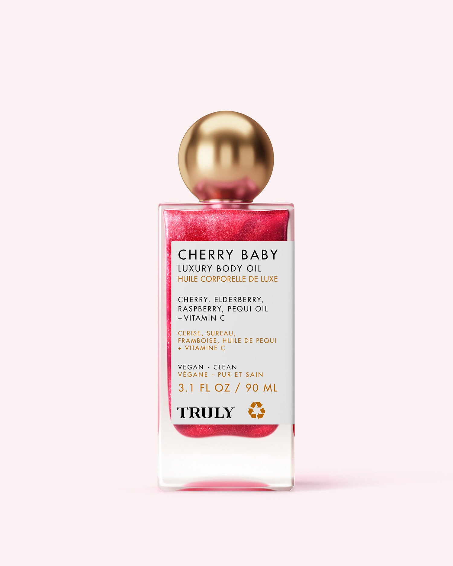 A rectangular Truly Beauty Cherry Baby Luxury Body Oil bottle with a shiny gold cap, pink shimmery oil, and a white label listing ingredients and benefits in English and French. Set against a light pink background to show its radiant shimmer.