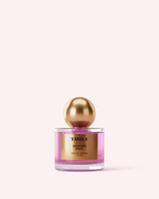 A bottle from Truly Beauty’s The Perfumers Set features pink liquid, a gold cap, and a gold label against a light pink backdrop—your next signature scent among Truly Beauty’s intoxicating fragrances.
