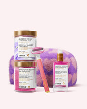 The Truly Beauty Ultimate Smooth Legend Shave Set includes Glazed Donut and Smooth Legend skincare, two jars, a bottle, a razor, and a purple-and-pink fuzzy case—all arranged on a light pink background.