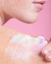 A close-up of someone applying Truly Beautys Unicorn Fruit Duo, a pastel-colored whipped body butter, to their freckled shoulder against a pink background.