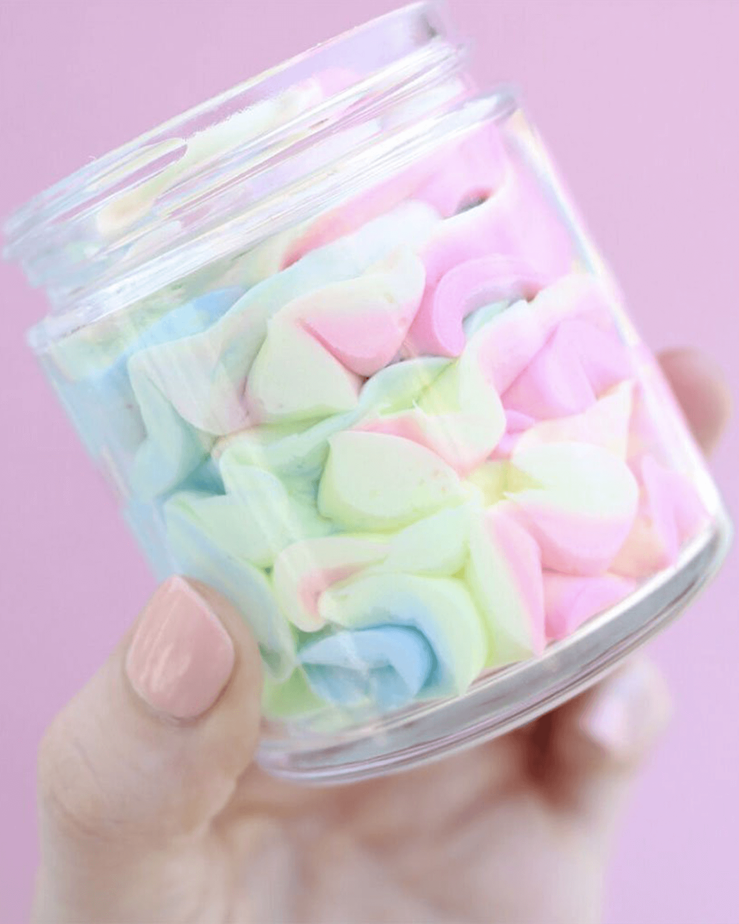 A hand with light pink nail polish holds a clear jar of Truly Beautys Unicorn Fruit Duo against a pale pink background, evoking the sweet allure of pastel-colored, cotton candy-scented treats.
