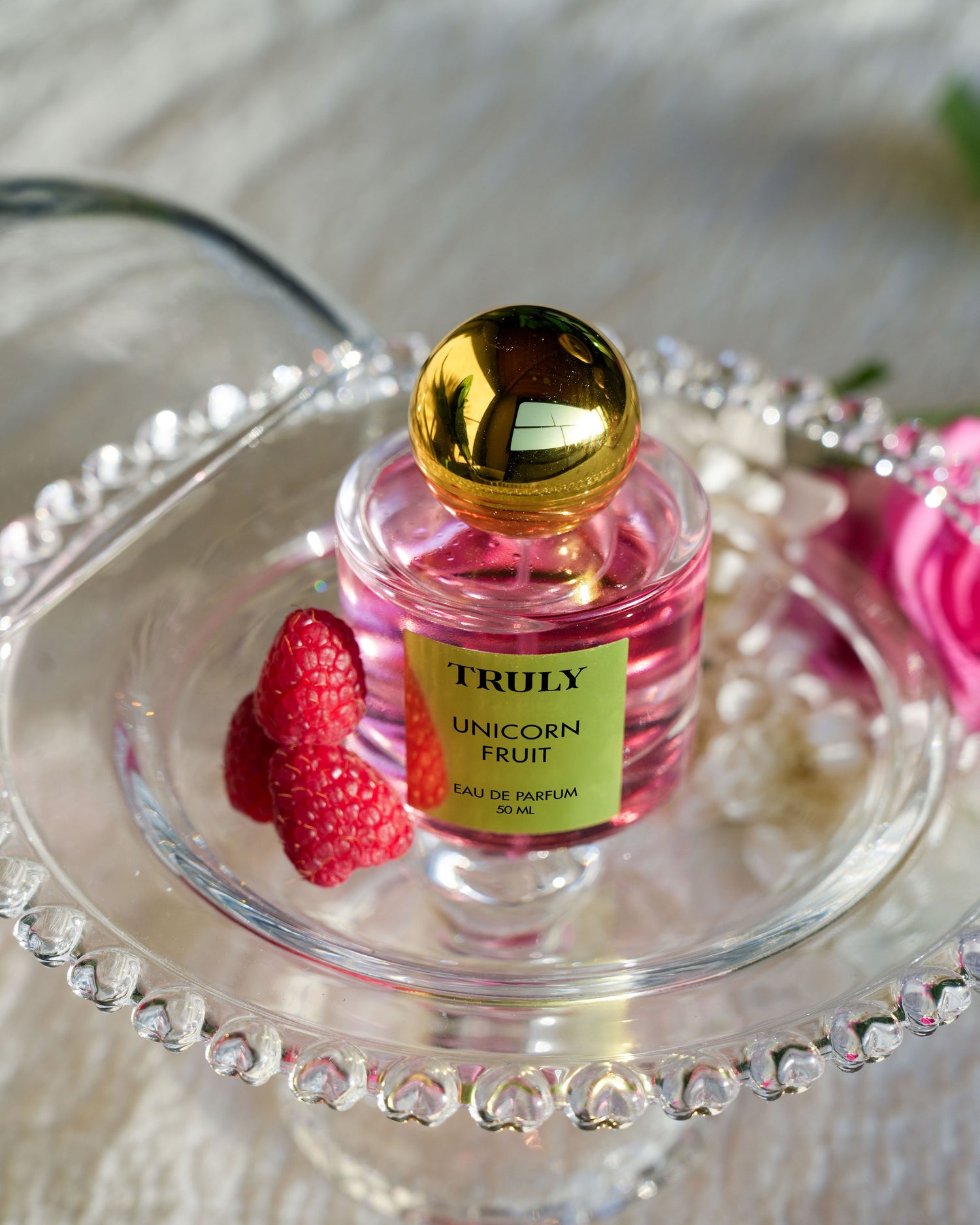 A bottle of Truly Beauty’s Unicorn Fruit Eau De Parfum with a gold cap rests in a glass dish beside raspberries and white flowers, evoking the sweet, fruity, and feminine scent of Unicorn Fruit Body Butter.