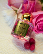 A pink bottle of Truly Beautys Unicorn Fruit Eau De Parfum with a gold cap rests among pink roses, white flowers, and two raspberries—reflecting its fruity, feminine scent inspired by the popular Unicorn Fruit Body Butter.