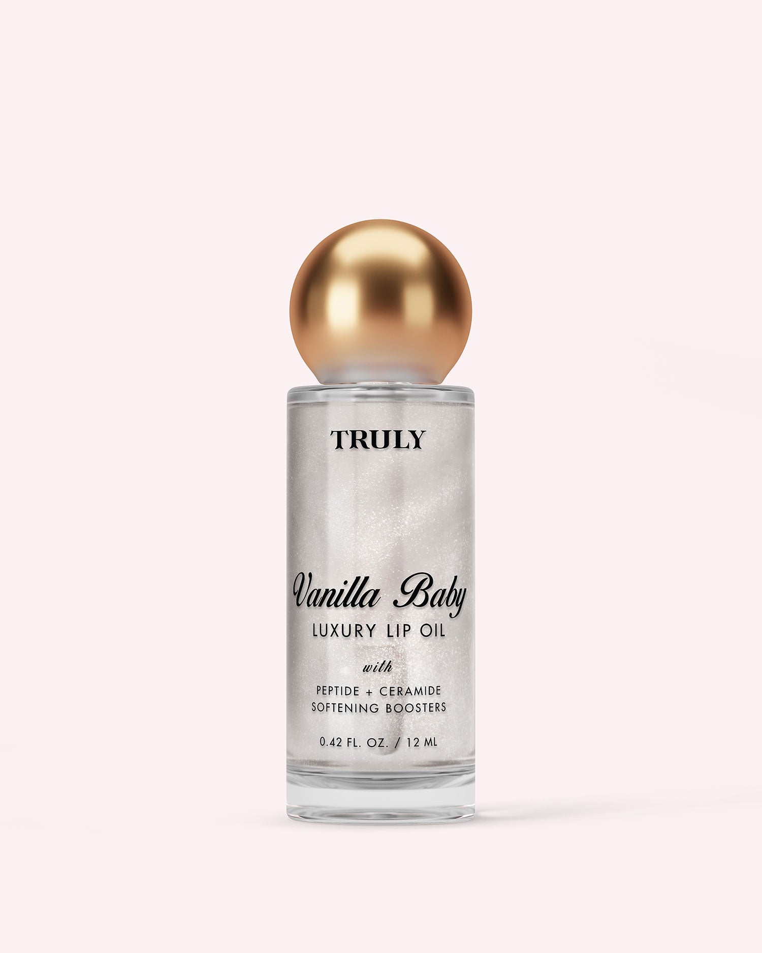 A clear bottle of Truly Luxury Body Oils by Vanilla Baby features a gold spherical cap, black text, and shimmering oil inside, set against a light pink background.