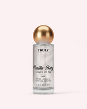 A clear bottle of Truly Luxury Body Oils by Vanilla Baby features a gold spherical cap, black text, and shimmering oil inside, set against a light pink background.