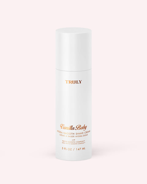 A white cylindrical bottle labeled Hydra-Smooth Shave Cream by Truly Beauty, with gold and peach text, sits on a light pink background. The bottle contains 5 fl oz (147 ml) of product.